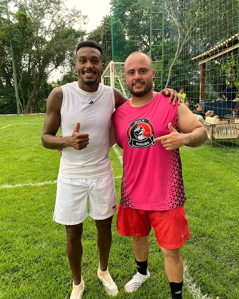 O Mateus de uniforme ao lado de Edenilson, num campo de futebol