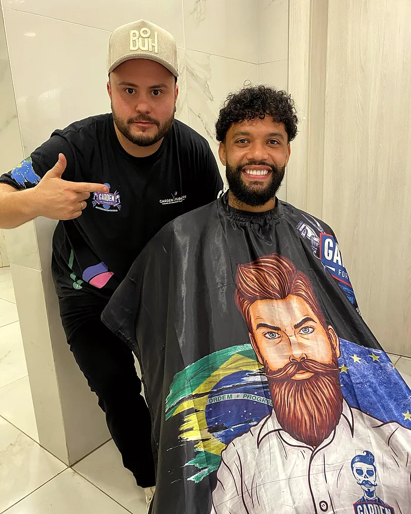 Otávio sentado na cadeira com a capa da barbearia, ao lado do Mateus